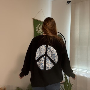 Puede incluir: Sudadera negra con un gran símbolo de la paz en la espalda. El símbolo de la paz es un diseño floral azul y blanco. La sudadera es de estilo casual.