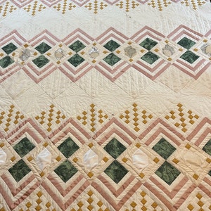 May include: A white quilt with a geometric pattern in shades of pink, green, and gold. The quilt is made up of squares and diamonds, and the pattern is repeated throughout the quilt.
