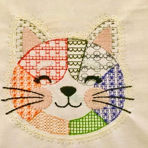 May include: Embroidered cat face with a patchwork design in shades of red, orange, green, blue, and white. The cat has black whiskers and a black nose. The design is on a cream-colored fabric and is framed with a delicate, cream-colored lace.