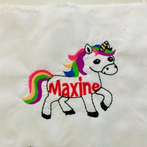 May include: A white fabric patch featuring an embroidered unicorn with a rainbow-coloured mane and tail. The unicorn has a gold-striped horn and black hooves. The name Maxine is embroidered in red thread across the body of the unicorn.