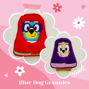 May include: Two embroidered patches featuring cartoon dog characters dressed as grannies. The larger patch shows a blue dog in a red hooded cloak with purple glasses. The smaller patch shows a tan dog in a purple hooded cloak. Text reads Blue Dog Grannies.