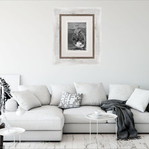 May include: A white sectional sofa with grey throw pillows and a grey throw blanket. A framed black and white photograph hangs on the wall above the sofa. A small white table with a cup and saucer sits in front of the sofa. A potted plant is on the right side of the image.