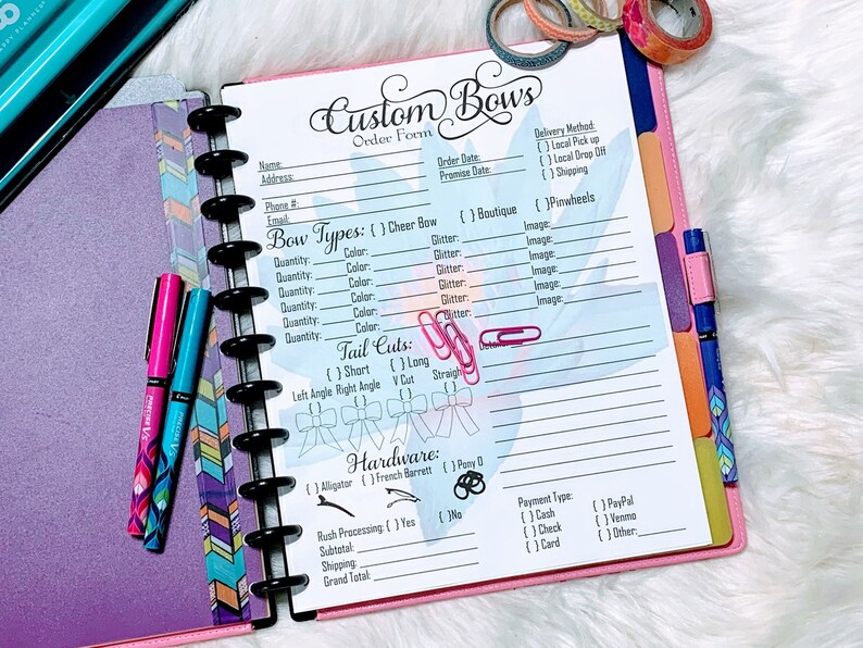 May include: A pink and white planner with a custom bow order form. The form includes sections for bow types, tail cuts, hardware, rush processing, subtotal, shipping, and grand total. The form also includes sections for delivery method, payment type, and other details.