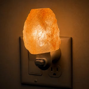 May include: A plug-in night light featuring a rough-cut, translucent orange Himalayan salt rock. The rock is illuminated from within, casting a warm, soft glow. It is attached to a white plastic base with a small toggle switch, plugged into a wall socket.