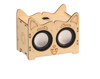 May include: A wooden speaker with a cat face design. The speaker has two round speakers and a small switch on the top. The speaker is made of light wood and has a natural finish.