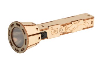 May include: A wooden toy flashlight with a clear lens and a black button. The flashlight is made of light-colored wood and features etched designs, including a battery symbol. The design is reminiscent of a DIY project or a craft kit.