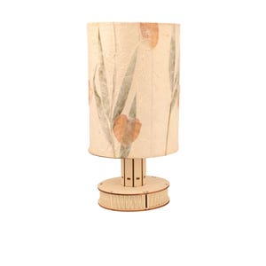 Könnte beinhalten: Eine Tischlampe mit einem cremefarbenen Lampenschirm, der mit gepressten Blumen und Blättern verziert ist. Der Lampenfuß ist aus Holz und hat eine zylindrische Form.