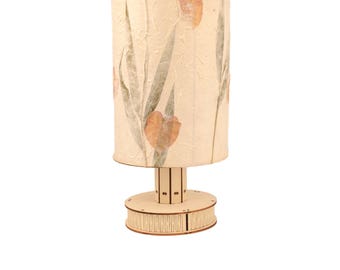 May include: A table lamp with a cream-colored shade featuring pressed flowers and leaves. The lamp base is made of wood and has a cylindrical shape.