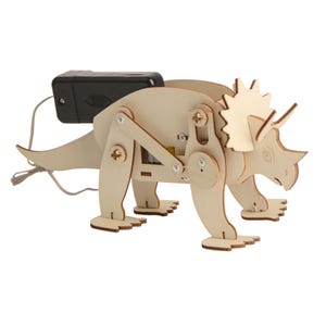 May include: A wooden Triceratops model kit with a battery pack attached. The dinosaur is constructed from light-colored wood and features articulated legs and a detailed head with horns. The kit includes small screws and a wire.