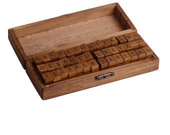 May include: A wooden box with a hinged lid, filled with small wooden letter and number blocks. The blocks are arranged in rows and columns, displaying the alphabet, numbers, and symbols. The box is made of light brown wood and has a simple, rustic design.