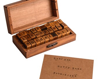 May include: A wooden box containing a set of alphabet stamps, with a hinged lid and metal clasp. The stamps are arranged in rows. A brown card with the text "Dear Ann, Merry Xmas, From: Alice" is next to the box.
