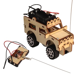 Puede incluir: Un kit de vehículo todoterreno de control remoto de madera para construir. El vehículo tiene ruedas negras, un compartimento para la batería y una barra de luces. El mando a distancia tiene una placa de circuito y una antena.