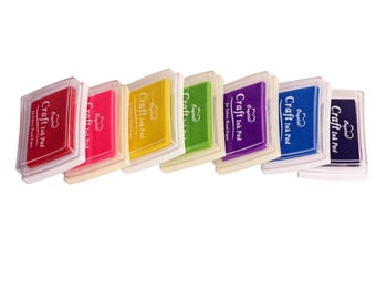 May include: A row of seven colorful craft ink pads in red, pink, yellow, green, purple, blue, and navy. Each pad has a clear plastic lid and a white base. The text "Craft Ink Pad" is visible on each pad.