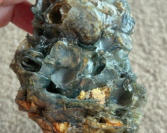 May include: A raw, uncut gemstone with a layered, translucent appearance. The stone features shades of gray, brown, and hints of orange. The rough texture and natural form suggest a unique, organic piece, possibly for collection or display.