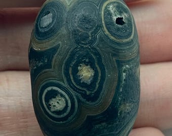 May include: An oval-shaped stone bead featuring concentric circular patterns in shades of dark blue, charcoal gray, and earthy brown. The surface displays natural textured inclusions and small crater-like markings within the layered mineral rings.