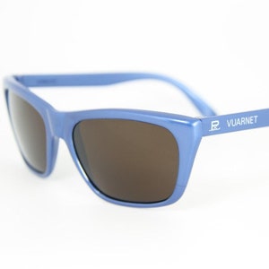 May include: Blue sunglasses with brown lenses. The sunglasses have a rectangular frame and the brand name "VUARNET" is printed on the arm. The sunglasses are set against a white background.