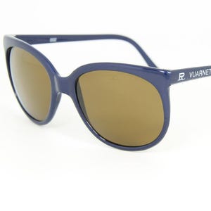 May include: Dark blue sunglasses with brown lenses. The sunglasses have a rounded frame and the brand name "VUARNET" is printed on the temple. The sunglasses are set against a white background.