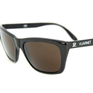 May include: Black sunglasses with brown lenses. The sunglasses have a rectangular frame and the brand name "VUARNET" is printed in white on the arm. The sunglasses are set against a white background.