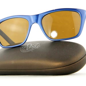 May include: Blue sunglasses with brown lenses rest on a dark brown case. The sunglasses have a rectangular shape with a blue frame and temples. The case has a rounded shape and a logo.