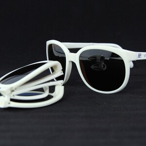 May include: White sunglasses with black lenses, one pair open and the other folded. The sunglasses have a rounded frame and the brand name "VUARNET" is printed on the arm. The folded pair shows the folding mechanism.