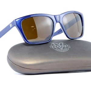 May include: Blue sunglasses with brown lenses rest on a gray case. The sunglasses have a square frame and a small logo on the temple. The case has the brand name "Vuarnet Sunglasses" printed on it.