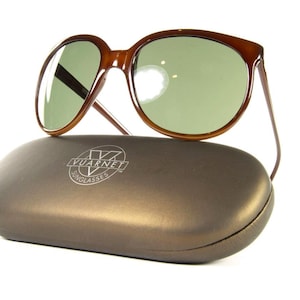 May include: Brown sunglasses with green lenses rest on a brown case. The sunglasses have a classic design with a thick frame. The case has the brand name "Vuarnet Sunglasses" printed on it.
