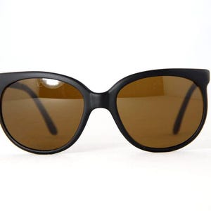 May include: Black sunglasses with brown lenses. The sunglasses have a rounded square shape and a matte finish. The lenses are a warm brown color, and the frames are a solid black. The sunglasses are set against a white background.