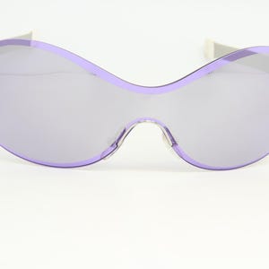 Puede incluir: Gafas de sol grandes, sin montura, con una forma curva distintiva. Las lentes son de color lavanda suave, y la montura es del mismo tono. Las patillas plateadas añaden contraste. Las gafas se presentan sobre un fondo blanco liso.
