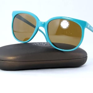May include: Teal sunglasses with brown lenses rest on a dark brown case. The sunglasses have a thick frame and the brand name is printed on the arm. The case is oval-shaped and has a smooth surface.