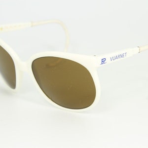 May include: White sunglasses with brown lenses. The sunglasses have a curved frame and ear pieces. The text "VUARNET" is printed in blue on the side of the frame.