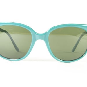 May include: A pair of turquoise sunglasses with a rounded frame and dark lenses. The sunglasses have a classic design, suitable for everyday wear. The frame is a solid, opaque color.