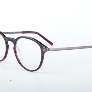 Puede incluir: Gafas de montura redonda en negro y burdeos. Las monturas tienen un diseño moderno con varillas metálicas y detalles burdeos. Las gafas se muestran sobre un fondo blanco.