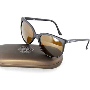 May include: Black sunglasses with brown lenses rest on a brown case. The sunglasses have a sleek, modern design with a matte finish. The case has a logo and the words "Vuarnet Sunglasses" printed on it. The sunglasses are a classic accessory.