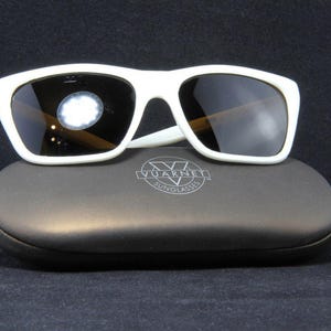 May include: White sunglasses with dark lenses rest on a dark gray case. The sunglasses have a thick, rectangular frame. The case has the brand name "Vuarnet Sunglasses" printed on it. The background is black.