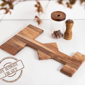 May include: A wooden serving board shaped like a T with a unique design. The board is made of light brown wood with a natural grain pattern. The board is perfect for serving cheese, crackers, and other appetizers. The text "THE ULTIMATE FAN GIFT!" is stamped on the board.