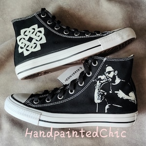 May include: A pair of black high-top Converse sneakers with custom hand-painted designs. One shoe features a white Celtic knot design, while the other shoe features a black and white silhouette of a person singing into a microphone. The shoes are labeled "HandpaintedChic".