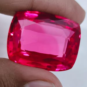 May include: A large, faceted, cushion-cut gemstone in a vibrant, saturated pink hue. The gem is held between fingers, showcasing its clarity and brilliance. The cut of the stone creates multiple reflective surfaces, enhancing its sparkle.