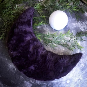 May include: A plush purple crescent moon shaped pillow with a white sphere in the background.