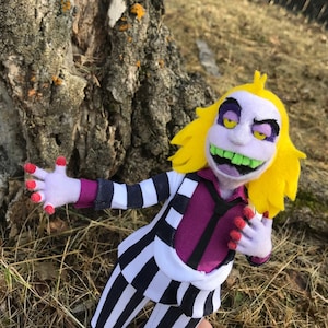 May include: A felt doll of a character with yellow hair, a purple shirt, and a black and white striped suit. The doll has a wide grin with green teeth and large, yellow eyes.