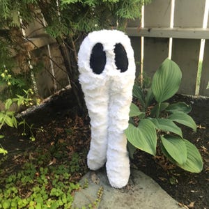 May include: White plush toy shaped like a pair of legs with black eyes. The toy is standing on a stone surface with green plants in the background.
