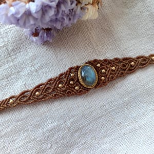 Könnte beinhalten: Ein braunes Makramee-Armband mit einem goldenen, ovalen Labradorit-Edelstein in der Mitte. Das Armband ist mit kleinen goldenen Perlen gewebt.