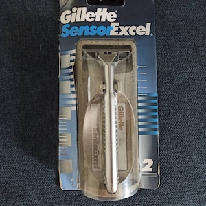 May include: Gillette SensorExcel razor in its original packaging. The packaging is primarily blue with white and silver text. The razor is silver and encased in a clear plastic. The packaging indicates that there are two razors included.