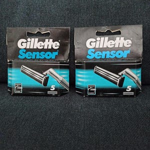 May include: Two packages of Gillette Sensor razors. Each package is black with the Gillette logo and the word "Sensor" in blue. The packaging shows a silver razor with five blades. The packages are on a dark gray surface.