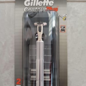 Pode incluir: Uma nova lâmina de barbear Gillette Contour Plus na embalagem original. A lâmina tem um cabo prateado e uma cabeça preta. A embalagem é preta e cinza com o logotipo da Gillette e o número "2" em laranja.