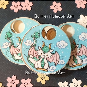 May include: Three round, blue and white painted wooden ornaments with a dragon design. Each ornament has a small hole in the center. The dragon is green, white, and pink with a cloud background. The text "Butterflymoon.Art" is printed on the ornaments.