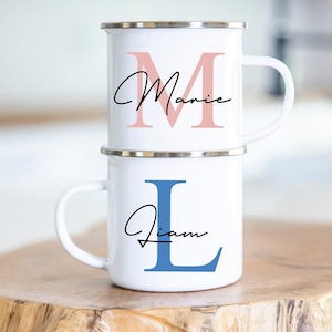 Könnte beinhalten: Zwei weiße Emailletassen mit silbernem Rand. Die obere Tasse hat ein rosafarbenes "M" mit dem Namen "Marie" in schwarzer Schrift. Die untere Tasse hat ein blaues "L" mit dem Namen "Liam" in schwarzer Schrift.