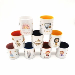 Könnte beinhalten: Neun weiße Keramikbecher mit bunten Designs und aufgedruckten Namen. Die Becher sind pyramidenförmig gestapelt. Die Designs beinhalten einen Regenbogen, einen LKW, einen Löwen, eine Meerjungfrau, einen Roboter, einen Ritter, einen Mantarochen, einen Feuerwehrmann und eine Prinzessin.