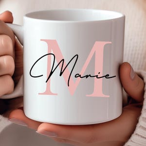 Könnte beinhalten: Weiße Keramiktasse mit einem großen rosa Buchstaben "M" und dem Namen "Mamie" darunter in Kursivschrift.