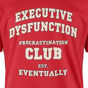 May include: A red t-shirt featuring the text "EXECUTIVE DYSFUNCTION PROCRATINATION CLUB EST. EVENTUALLY" in a white, vintage font. The shirt is a solid red colour and has short sleeves.
