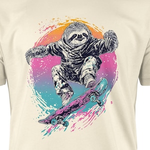 May include: Cream-colored t-shirt featuring a graphic of a sloth skateboarding. The sloth is depicted in black and white, with a colorful splash of pink, blue, and orange behind it. The skateboard is blue and pink.